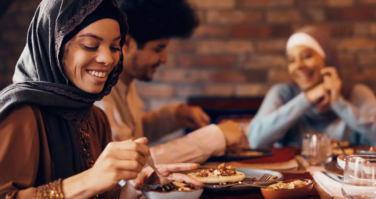 A woman with a headscarf smiles as she enjoys a meal at a table with two other people. Plates of food are visible in front of them, creating a warm and inviting atmosphere focused on sharing and companionship.