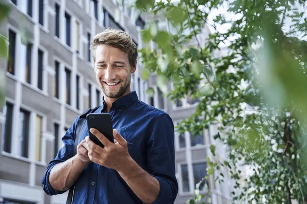 A smiling man with light hair stands outdoors, holding a smartphone and looking at the screen. He is dressed in a blue shirt, surrounded by greenery and urban buildings, appearing engaged and happy with whatever he is viewing on his phone.