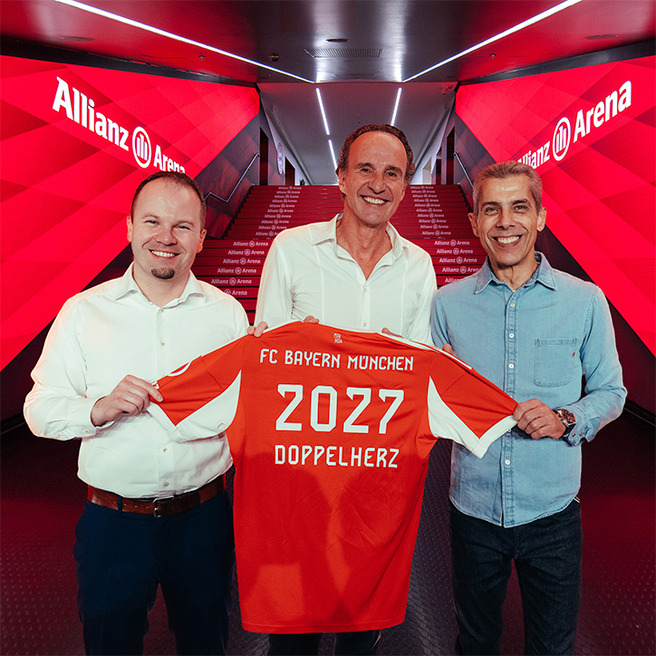 Trois personnes se tiennent dans un couloir de l'Allianz Arena, tenant un maillot de foot orange du FC Bayern Munich. Le maillot affiche "2027" et "DOPPELHERZ" au dos. L'ambiance est festive, suggérant un événement important pour le club.