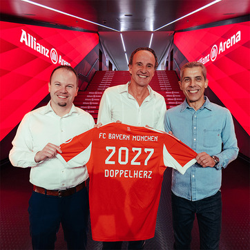 Trois personnes se tiennent dans un couloir de l'Allianz Arena, tenant un maillot de foot orange du FC Bayern Munich. Le maillot affiche "2027" et "DOPPELHERZ" au dos. L'ambiance est festive, suggérant un événement important pour le club.