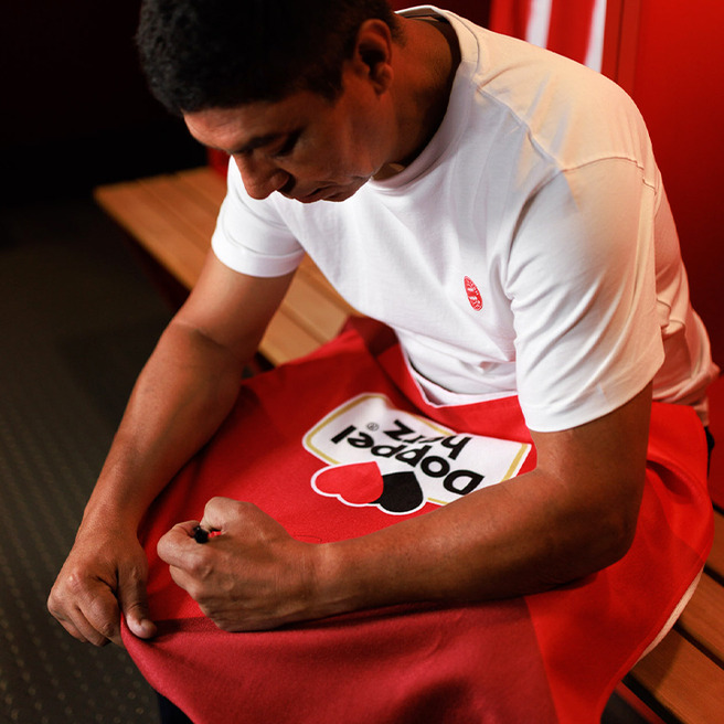 Un homme assis dans un vestiaire, portant un t-shirt blanc et un pantalon rouge. Il ajuste une serviette rouge avec un design coloré au centre. L'environnement est simple, avec des bancs en bois et un fond rouge.