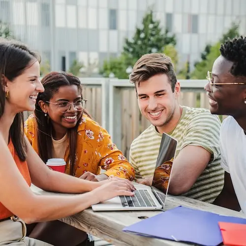 Quatre jeunes adultes se réunissent autour d'un ordinateur portable sur une table extérieure. Ils sourient et échangent des idées, créant une atmosphère de collaboration et de camaraderie. L'un d'eux tient une tasse de café. L'arrière-plan présente des bâtiments modernes.