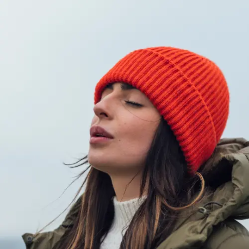 Une femme portant un bonnet orange regarde vers le haut, les yeux fermés, avec un air apaisé. Elle est vêtue d'un manteau olive et se trouve en extérieur, suggérant une ambiance tranquille et contemplative.