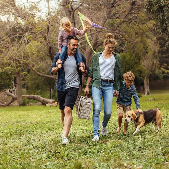 Une famille se promène dans un parc verdoyant. Un homme porte une petite fille sur ses épaules, tandis qu'une femme tient la main d'un garçon qui marche à côté d'un chien. Ils semblent joyeux, partageant un moment convivial en plein air.