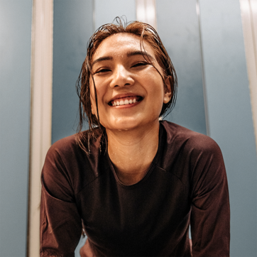 Une femme souriante avec les cheveux mouillés porte un haut noir. Elle se penche légèrement en avant, semblant joyeuse après une séance d'entraînement ou d'activité physique dans un environnement intérieur. Les murs derrière elle sont de couleur bleue.