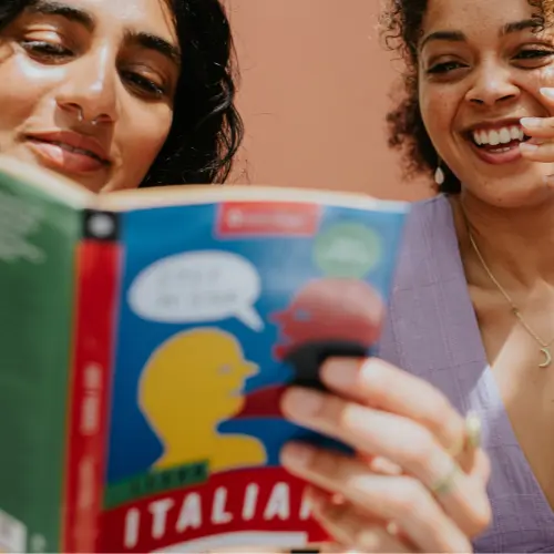 Deux femmes souriantes regardent un livre de conversation en italien. L'une tient le livre ouvert, montrant une couverture colorée avec des silhouettes. L'ambiance est joyeuse et amicale, mettant en avant le partage de l'apprentissage d'une nouvelle langue.
