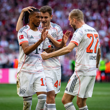 Des joueurs du Bayern Munich célèbrent un but. L'un d'eux, au maillot numéro 14, est entouré de ses coéquipiers qui expriment leur joie. L'arrière-plan montre une foule enthousiaste, typique d'un match de football.