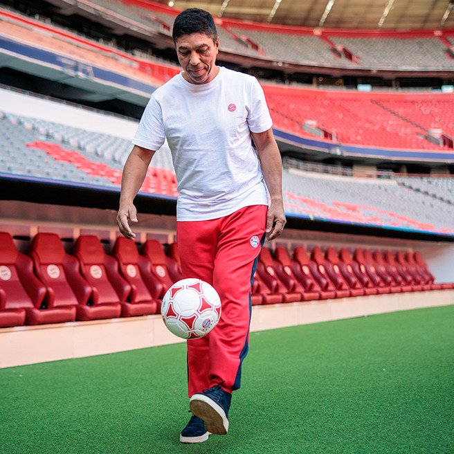 Un homme souriant porte un t-shirt blanc et un pantalon rouge, dribblant un ballon de football dans un stade vide. Les sièges rouges sont visibles en arrière-plan. L'homme semble apprécier l'activité sur le terrain.