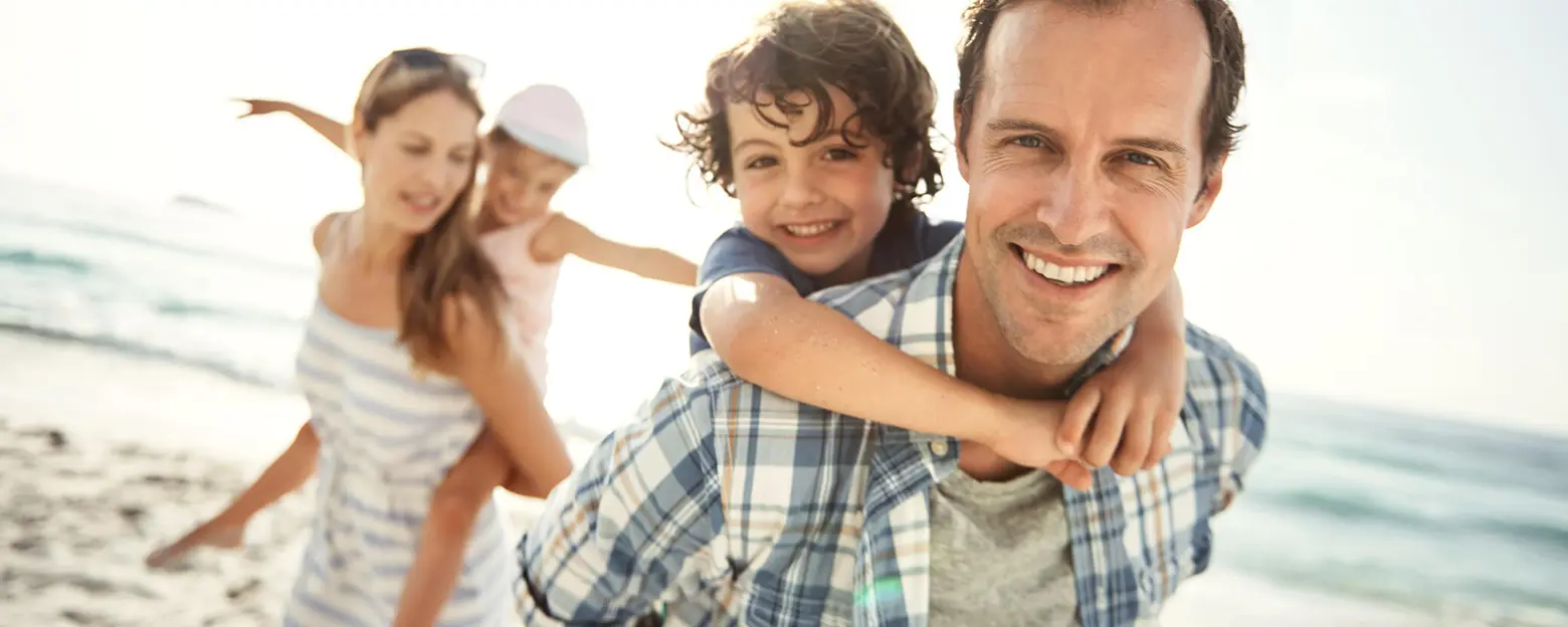 Une famille profite d'une journée à la plage. Un père souriant porte un garçon sur son dos, tandis que deux filles jouent à proximité. L'ambiance est joyeuse, avec un éclairage ensoleillé et des vagues en arrière-plan.