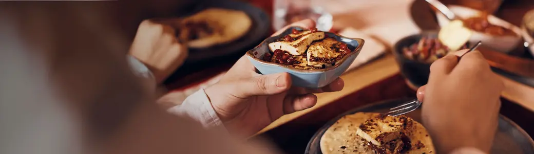 لقطة تُظهر يدًا تحمل وعاء صغير يحتوي على طبق من الطعام، بينما تُرى أطباق أخرى على الطاولة. الشخص يبدو مستعدًا لتناول الطعام باستخدام شوكة. تركز الصورة على تجربة تناول الوجبة مع أجواء دافئة ومريحة.